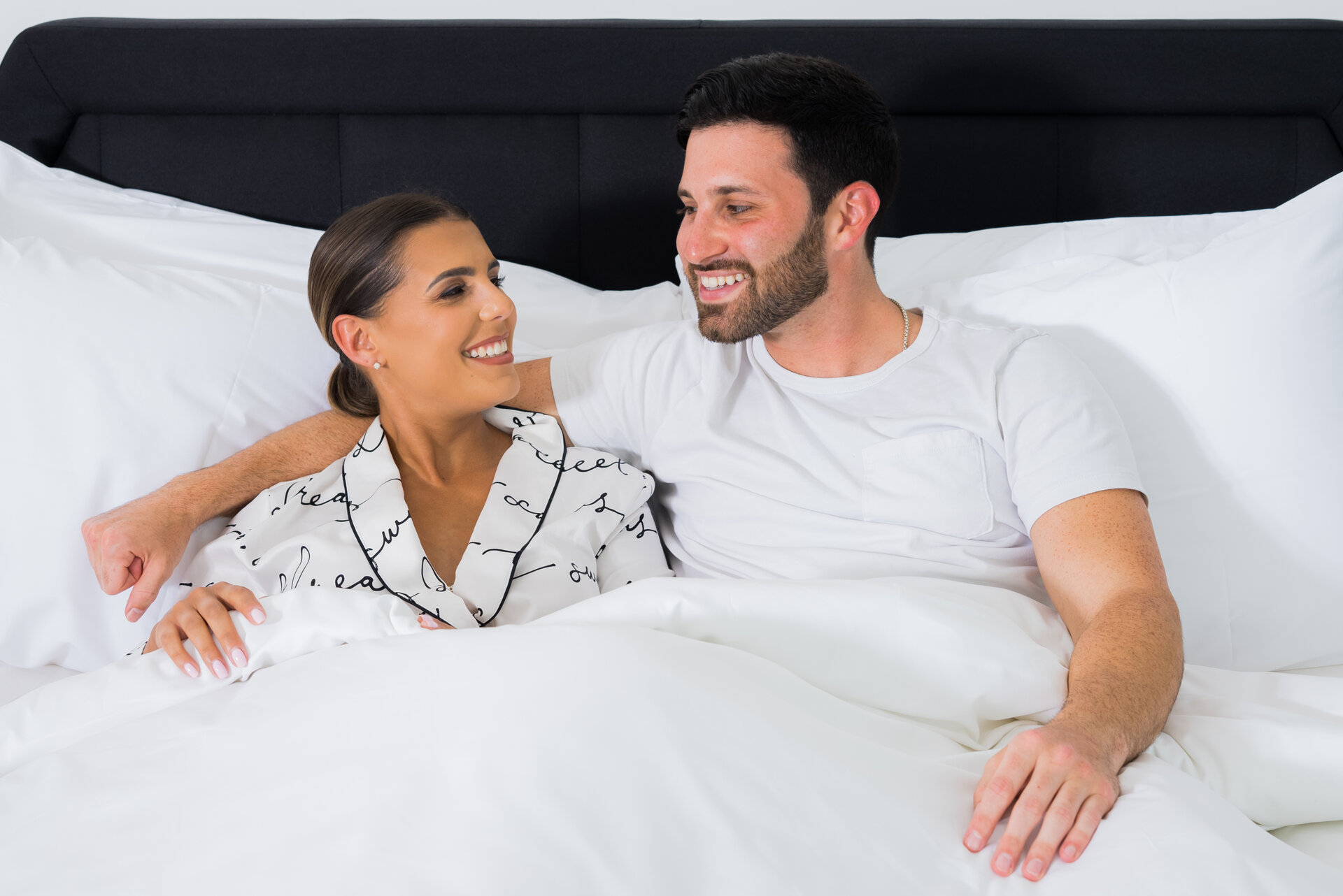 A man and woman in bed, smiling at each other.