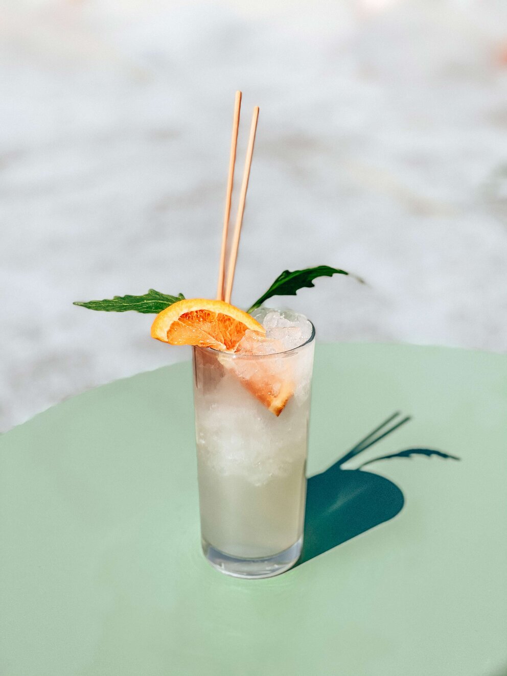 a tall glass of a clear liquid with ice, garnished with an orange slice and mint leaves, and two straws, on a green table
